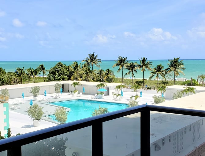 Zona de piscina de un complejo turístico con agua turquesa, sombrillas azules y palmeras, con vistas al mar tropical y la playa